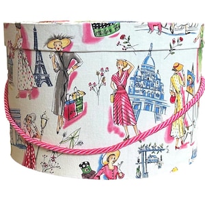 Può includere: Una cappelliera rotonda con sfondo bianco, con manico in corda rosa. La scatola è decorata con un motivo ripetuto di illustrazioni di donne in stile vintage, con monumenti come la Torre Eiffel.