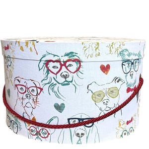 Può includere: Una scatola regalo rotonda bianca con un motivo ripetuto di cani con occhiali e cuori. La scatola ha un manico in corda rossa e un coperchio. I cani sono in vari colori e stili.