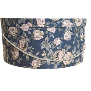 Puede incluir: Caja ovalada con estampado floral sobre fondo azul marino. Presenta un patrón repetido de rosas rosas y crema y hojas verdes. Tiene un asa de cuerda. Ideal para guardar o regalar.