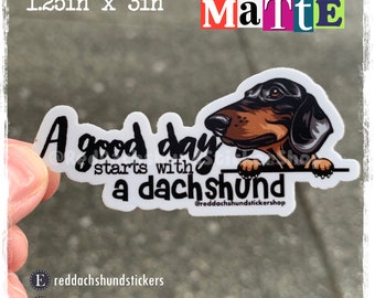 May include: A white sticker with a black and brown dachshund peeking over the top. The sticker says "A good day starts with a dachshund" and includes the social media handle "@reddachshundstickershop".