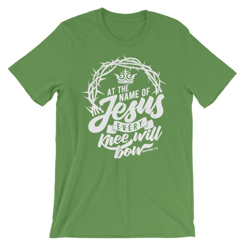 May include: Green t-shirt with a white graphic design featuring a crown of thorns, the words "At the name of Jesus every knee will bow", and the scripture reference "Philippians 2:10".