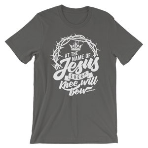May include: A dark gray t-shirt with a white graphic design. The graphic features a crown of thorns, a crown, and the text "At the name of Jesus every knee will bow Philippians 2:10".