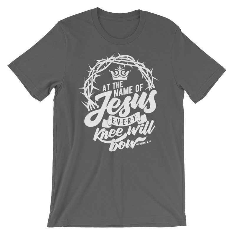 May include: A dark gray t-shirt with a white graphic design. The graphic features a crown of thorns, a crown, and the text "At the name of Jesus every knee will bow Philippians 2:10".