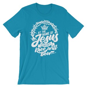May include: A turquoise blue t-shirt with a white graphic design. The graphic features a crown of thorns, a crown, and the text "At the name of Jesus every knee will bow Philippians 2:10".