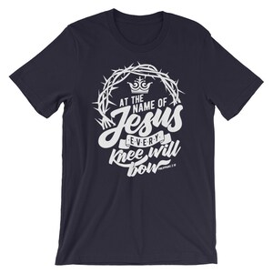 May include: Navy blue t-shirt with a white graphic design featuring a crown of thorns, the words "At the name of Jesus every knee will bow", and the scripture reference "Philippians 2:10".