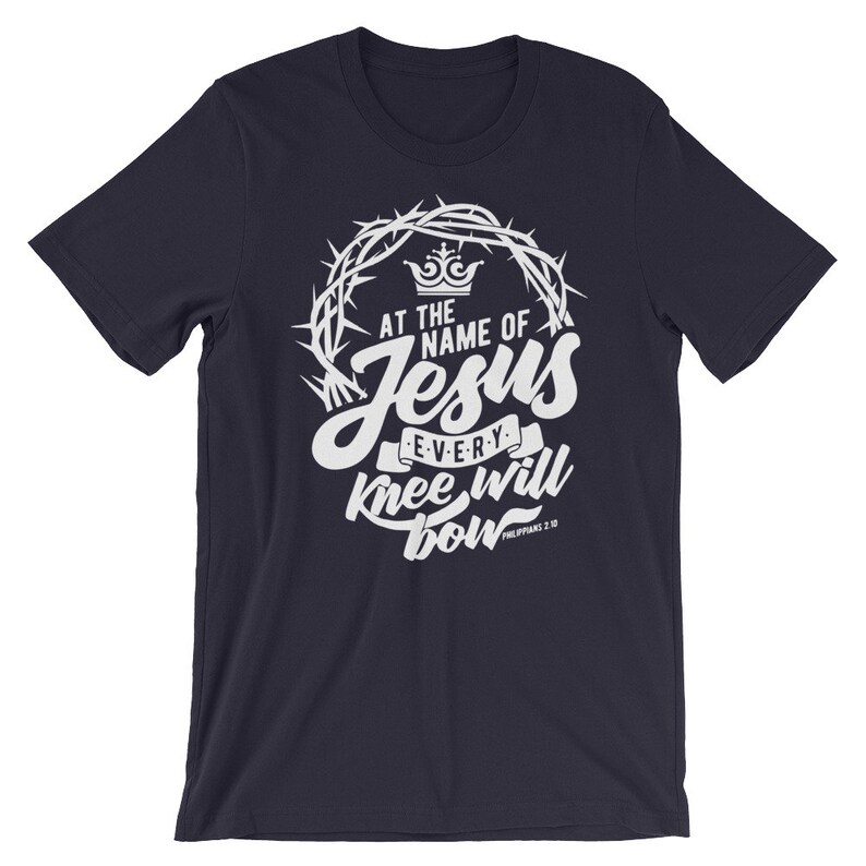May include: Navy blue t-shirt with a white graphic design featuring a crown of thorns, the words "At the name of Jesus every knee will bow", and the scripture reference "Philippians 2:10".