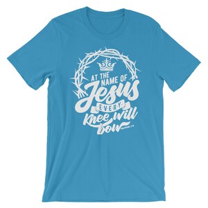 May include: A light blue t-shirt with a white graphic design. The graphic features a crown of thorns, a crown, and the text "At the name of Jesus every knee will bow Philippians 2:10".