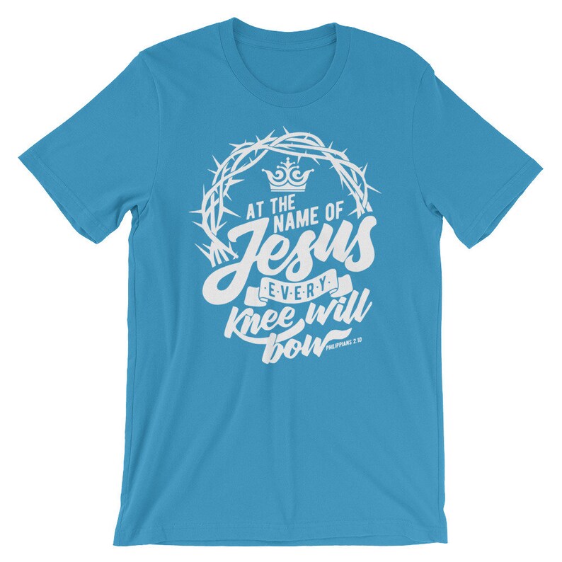 May include: A light blue t-shirt with a white graphic design. The graphic features a crown of thorns, a crown, and the text "At the name of Jesus every knee will bow Philippians 2:10".