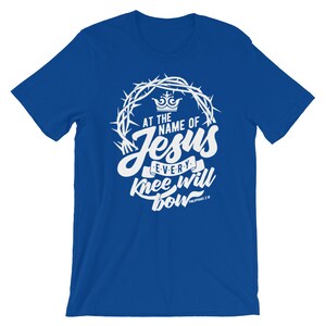 May include: A royal blue t-shirt with a white graphic design. The graphic features a crown, a crown of thorns, and the text "At the name of Jesus every knee will bow Philippians 2:10".