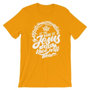 May include: A yellow t-shirt with a white graphic design. The graphic features a crown, thorns, and the words "At the name of Jesus every knee will bow" with the scripture reference "Philippians 2:10".