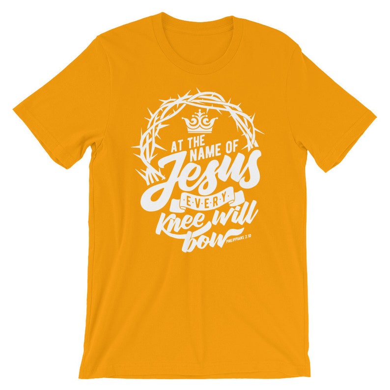 May include: A yellow t-shirt with a white graphic design. The graphic features a crown, thorns, and the words "At the name of Jesus every knee will bow" with the scripture reference "Philippians 2:10".