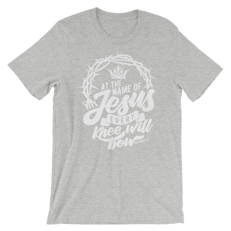 May include: Gray t-shirt with a white graphic design. The graphic features a crown, thorns, and the text "At the name of Jesus every knee will bow" with the scripture reference "Philippians 2:10".