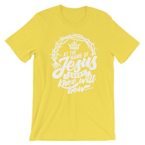 May include: Yellow short-sleeved t-shirt with a white graphic design. The graphic features a crown, thorns, and the text "At the name of Jesus every knee will bow" with the scripture reference "Philippians 2:10".