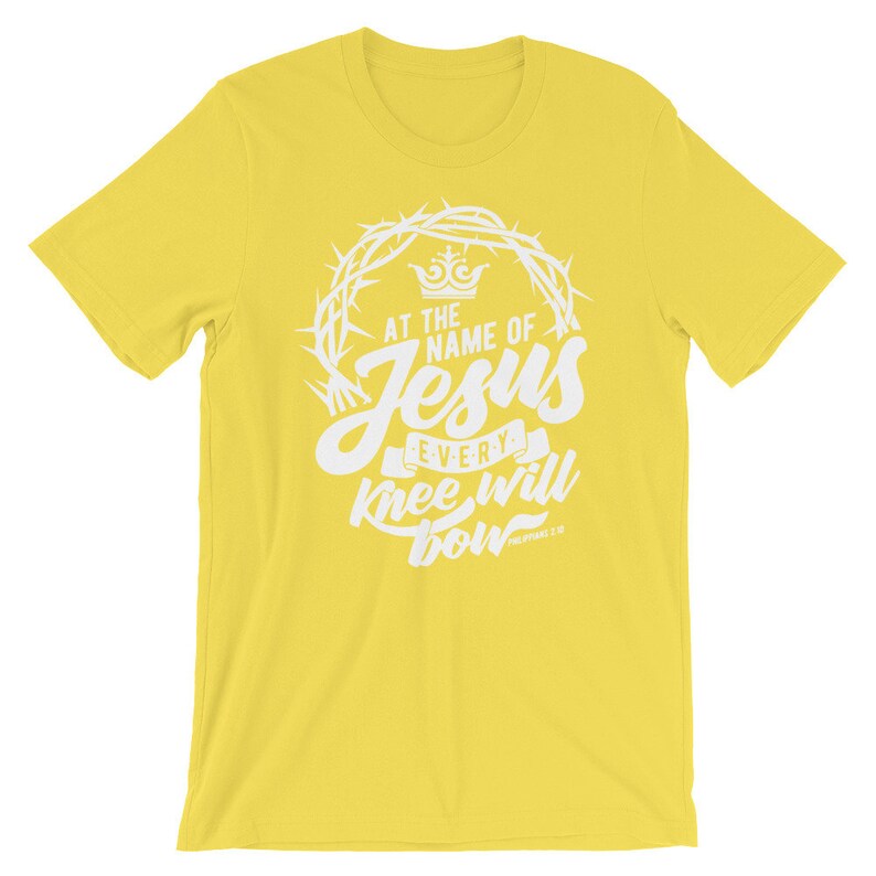 May include: Yellow short-sleeved t-shirt with a white graphic design. The graphic features a crown, thorns, and the text "At the name of Jesus every knee will bow" with the scripture reference "Philippians 2:10".