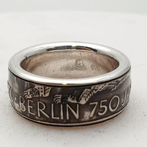 Op de afbeelding: Een zilveren ring met een ontwerp van een vogel en de woorden "BERLIN 750 JA" rond de band.