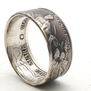 Pode incluir: Anel de prata feito com uma moeda alemã de 5 marcos. O anel apresenta um desenho de uma mulher segurando uma espada e um escudo com o texto "UND ALLE KAMMEN." ao redor da borda. O outro lado do anel tem o texto "5 MARK" e uma estrela.