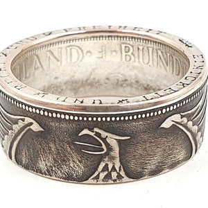 Pode incluir: Um anel de prata feito com uma moeda alemã de 5 Deutsche Mark. O anel apresenta um design de uma águia de duas cabeças com as palavras "Bundesrepublik Deutschland" ao redor da borda.