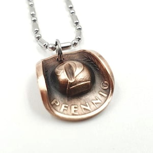 Könnte beinhalten: Eine silberne Kettenkette mit einem kupfernen Anhänger in Form eines Cowboyhuts. Der Anhänger ist eine umfunktionierte 2-Pfennig-Münze mit dem Text "2 PFENNIG" sichtbar.