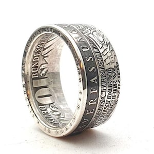 Op de afbeelding: Een zilveren ring met een ingelegde Duitse 10 Deutsche Mark-munt. De munt heeft de tekst "Bundesrepublik Deutschland" en "10 Deutsche Mark" rond de rand.