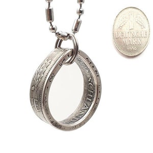 Puede incluir: Collar de plata con un colgante hecho de una moneda alemana Deutsche Mark de 1966. La moneda está montada en forma de anillo y la inscripción "DEUTSCHE MARK 1966" es visible en la moneda.