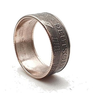 Pode incluir: Um anel de prata feito com uma moeda alemã de 1 Deutsche Mark. O anel apresenta a inscrição "1 DEUTSCHE MARK" e um design de águia estilizado.