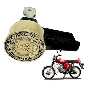 Könnte beinhalten: Ein Messing-Schlüssel mit einem runden Siegel mit dem Text "DEMOKRATISCHE REPUBLIK DEUTSCHE" und einem Kompass- und Hammer-Symbol. Der Schlüssel ist an einem schwarzen Metallgriff befestigt. Im Hintergrund ist ein rot-schwarzes Moped zu sehen.