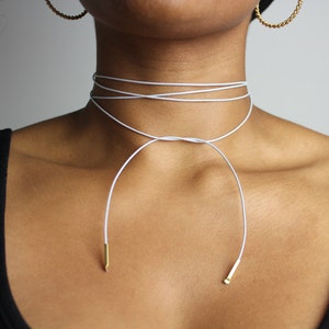 May include: A white leather choker necklace with gold accents. The necklace is tied in a knot at the front and has two long strands that hang down the chest.