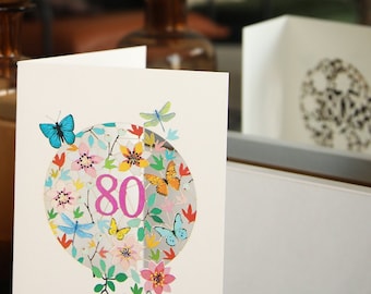 May include: A white greeting card featuring a central circular design filled with colorful illustrations of flowers, green leaves, butterflies, and dragonflies. The number 80 is printed in large pink numerals in the center of the circle.