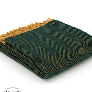 Può includere: Una coperta in lana verde e oro a spina di pesce, piegata con frange sui bordi. La coperta è in lana e ha un tocco morbido e lussuoso.