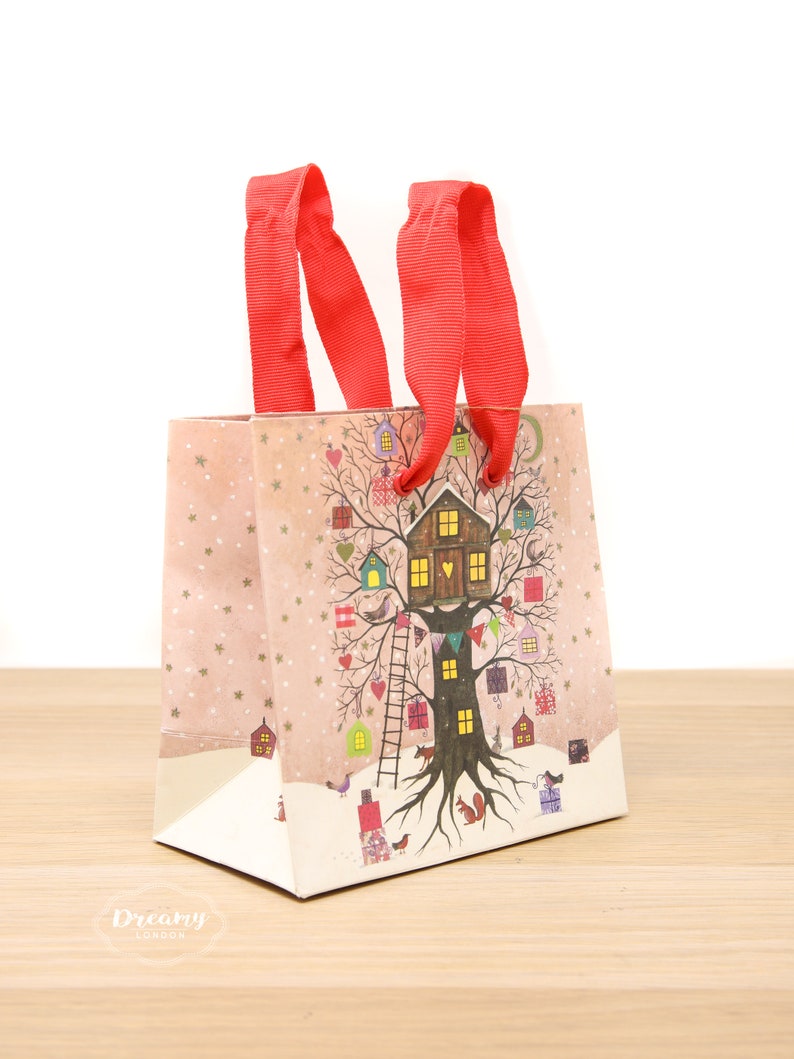 May include: A red and white gift bag with a winter scene featuring a tree house and a snowy landscape. The bag has red ribbon handles and the text "Dreamy London" is printed on the bottom.