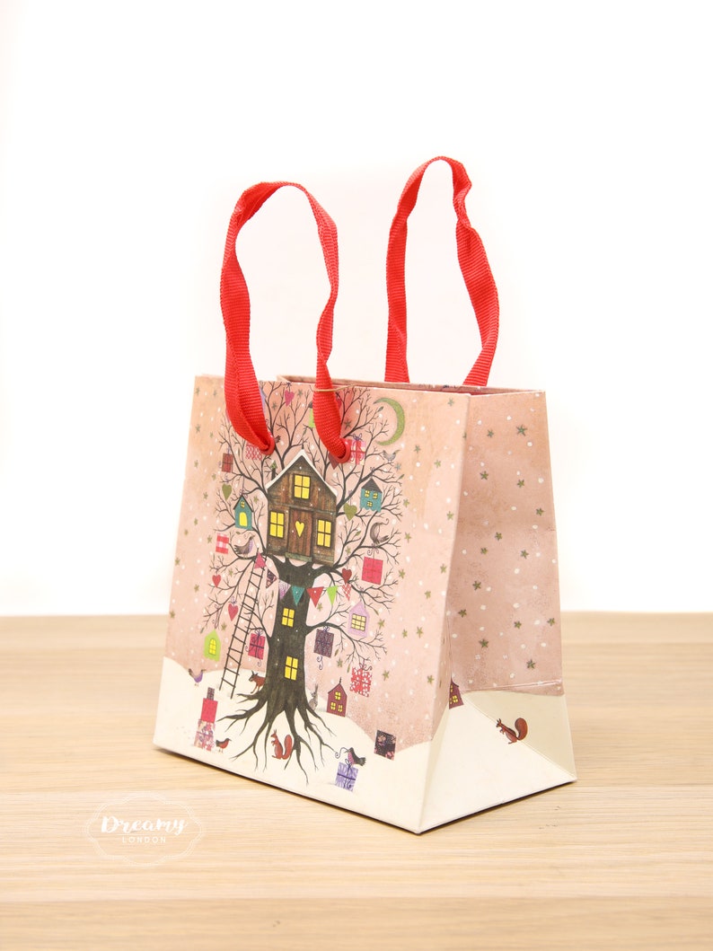 May include: A pink and white gift bag with red handles. The bag features a whimsical illustration of a tree house with a light on in the window. The tree is decorated with small houses, birds, and stars. The text "Dreamy London" is printed on the bottom of the bag.