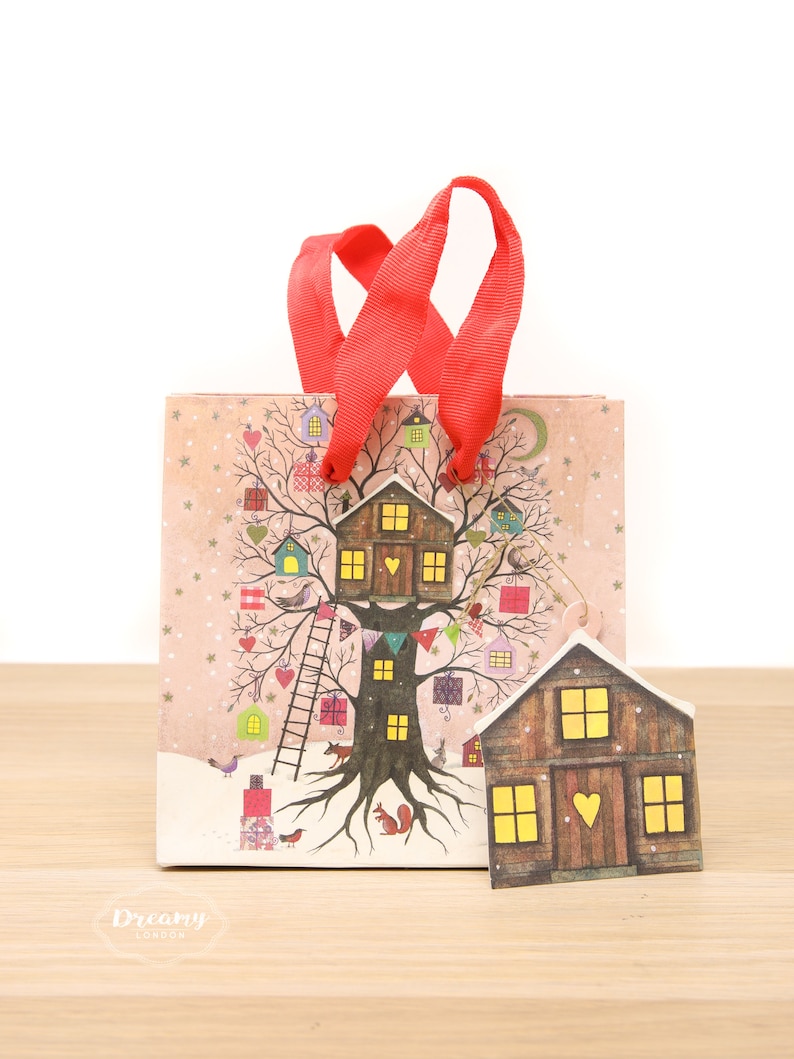 May include: A red gift bag with a white and pink treehouse design. The bag has a red ribbon handle and a small wooden house tag with a heart on the door. The bag has the text "Dreamy London" on the bottom.