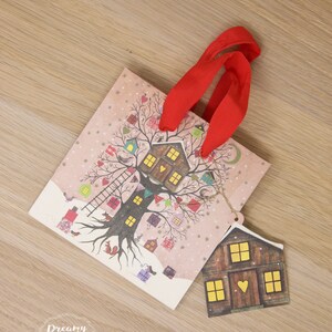 May include: A pink and white gift bag with a red ribbon handle. The bag features a whimsical illustration of a tree with houses and birds. A small brown house tag with yellow windows is attached to the bag.