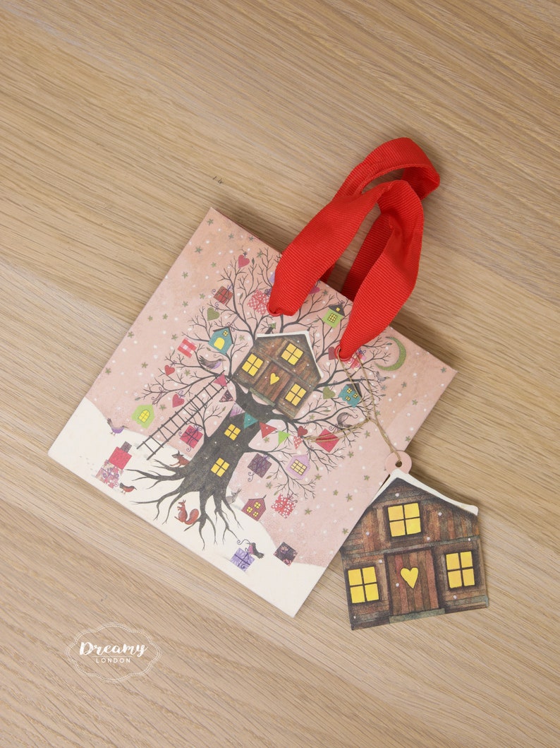 May include: A pink and white gift bag with a red ribbon handle. The bag features a whimsical illustration of a tree with houses and birds. A small brown house tag with yellow windows is attached to the bag.