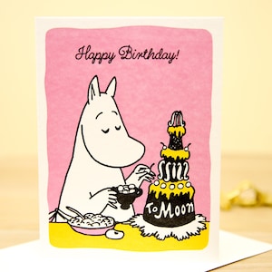 以下が含まれることがあります： 「To Moon」と書かれた黒と黄色のケーキと漫画のキャラクターが描かれたバースデーカード。「Happy Birthday!」の文字がピンク色の背景に黒い筆記体で書かれています。