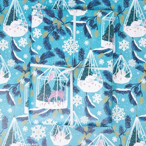 May include: Blue wrapping paper with a repeating pattern of white cages containing birds, deer, and snowflakes. The cages are suspended from a string and are surrounded by pine branches.