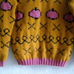 May include: A mustard yellow knitted sweater featuring a repeating pattern of pink pumpkin shapes, black stitched crosses, and black wavy lines. The garment has long sleeves and a ribbed pink hem at the waist and cuffs, constructed with a thick yarn texture.