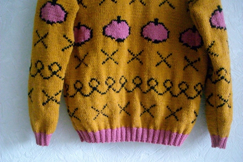 May include: A mustard yellow knitted sweater featuring a repeating pattern of pink pumpkin shapes, black stitched crosses, and black wavy lines. The garment has long sleeves and a ribbed pink hem at the waist and cuffs, constructed with a thick yarn texture.