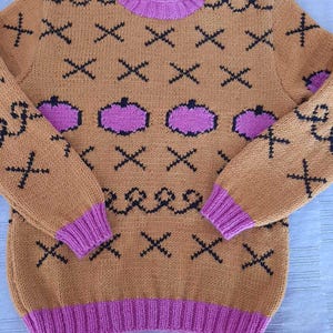 May include: A tan long-sleeve knit sweater featuring a repeating pattern of black X shapes and wavy lines, interspersed with purple pumpkin-shaped motifs. The garment includes a ribbed purple crew neckline, sleeve cuffs, and a wide ribbed purple hem.