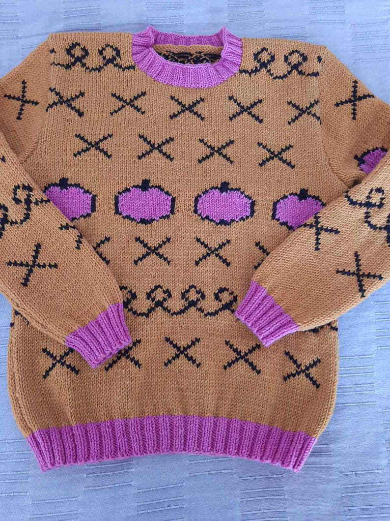 May include: A tan long-sleeve knit sweater featuring a repeating pattern of black X shapes and wavy lines, interspersed with purple pumpkin-shaped motifs. The garment includes a ribbed purple crew neckline, sleeve cuffs, and a wide ribbed purple hem.