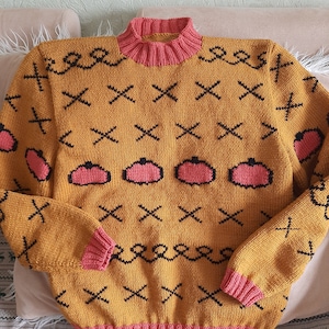 May include: A mustard yellow knit sweater featuring a pink ribbed mock neck, cuffs, and hem. The sweater is decorated with a repeating pattern of black stitched X shapes, black swirling lines, and pink pumpkin-shaped motifs with black outlines.