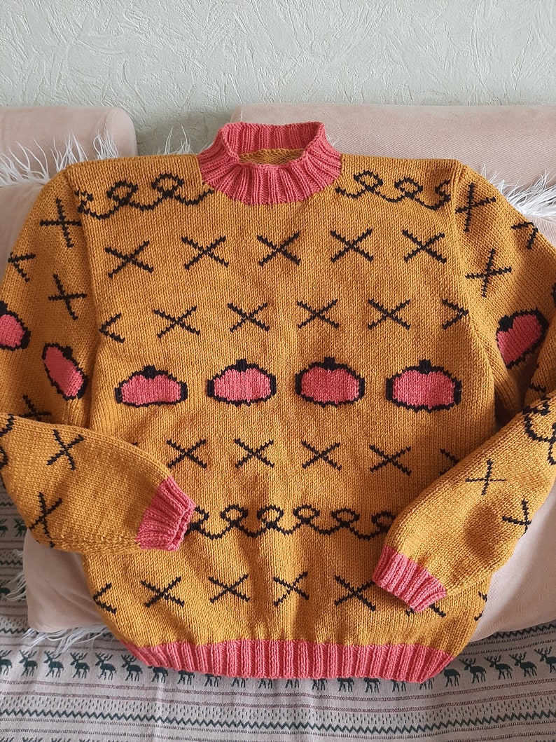 May include: A mustard yellow knit sweater featuring a pink ribbed mock neck, cuffs, and hem. The sweater is decorated with a repeating pattern of black stitched X shapes, black swirling lines, and pink pumpkin-shaped motifs with black outlines.