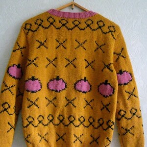 May include: A mustard yellow knit sweater featuring a repeating pattern of pink pumpkins, black X-shaped marks, and black wavy lines. The garment has a pink ribbed crew neckline, pink ribbed cuffs, and a pink ribbed hem, all displayed on a hanger.