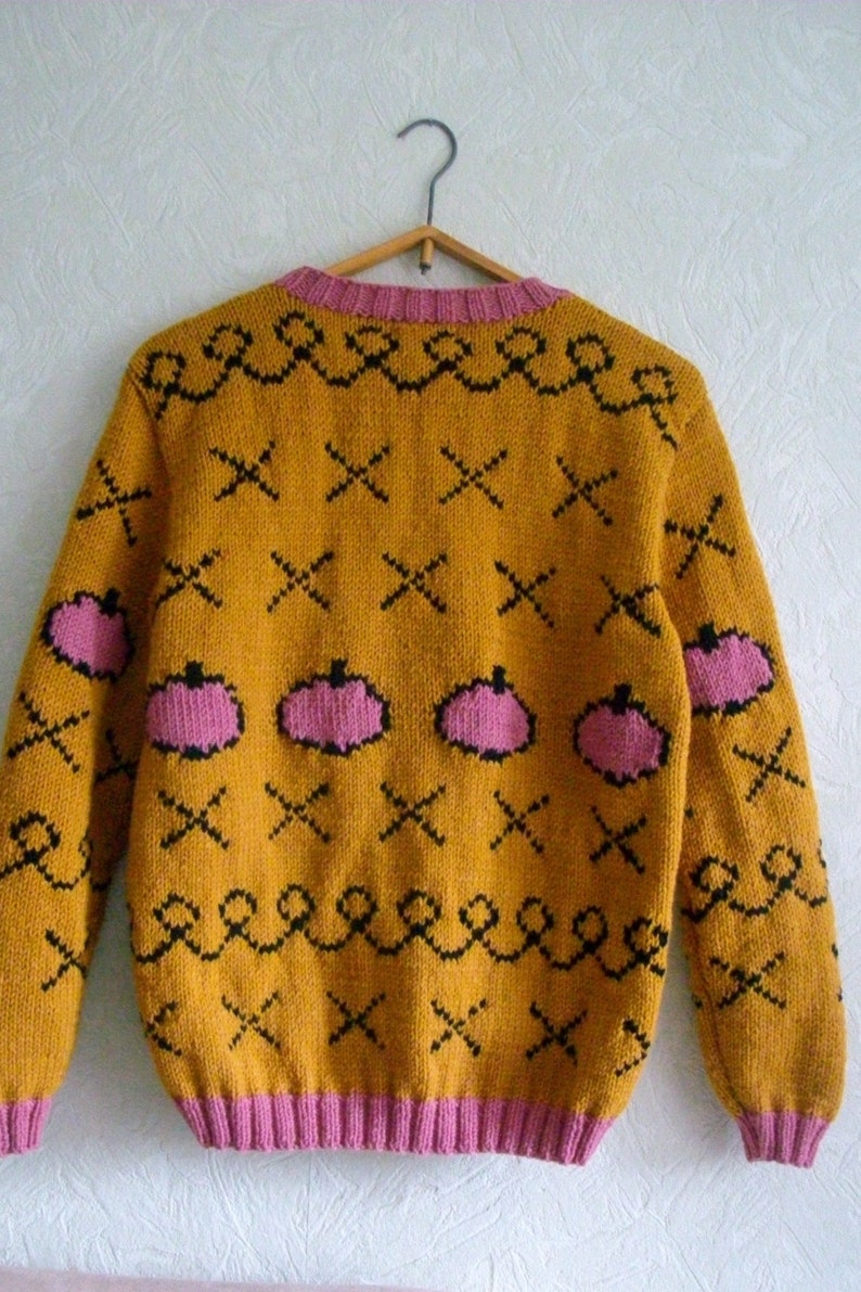 May include: A mustard yellow knit sweater featuring a repeating pattern of pink pumpkins, black X-shaped marks, and black wavy lines. The garment has a pink ribbed crew neckline, pink ribbed cuffs, and a pink ribbed hem, all displayed on a hanger.