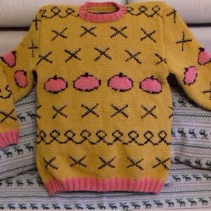 May include: A mustard yellow long-sleeve knit sweater featuring a repeating pattern of black stitched X shapes, black swirling lines, and pink pumpkin-shaped motifs. The garment has a pink ribbed crew neckline, pink ribbed cuffs, and a pink ribbed hem.