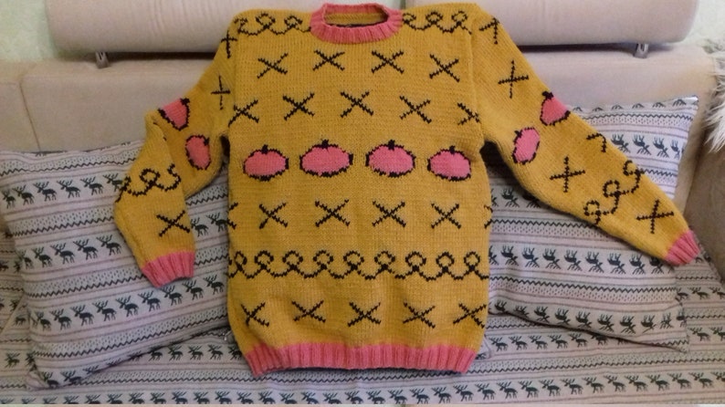May include: A mustard yellow long-sleeve knit sweater featuring a repeating pattern of black stitched X shapes, black swirling lines, and pink pumpkin-shaped motifs. The garment has a pink ribbed crew neckline, pink ribbed cuffs, and a pink ribbed hem.