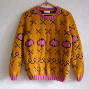 May include: A mustard yellow long-sleeve knit sweater featuring a pink ribbed crew neckline, cuffs, and hem. The sweater is decorated with a repeating pattern of black stitched X shapes, black wavy lines, and pink pumpkin-shaped motifs throughout.