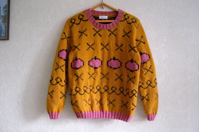 May include: A mustard yellow long-sleeve knit sweater featuring a pink ribbed crew neckline, cuffs, and hem. The sweater is decorated with a repeating pattern of black stitched X shapes, black wavy lines, and pink pumpkin-shaped motifs throughout.