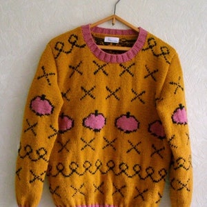 May include: A mustard yellow long-sleeve knit sweater featuring a repeating pattern of black cross stitches, black wavy lines, and pink pumpkin-shaped motifs. The garment has a pink ribbed crew neckline, cuffs, and hem, and a white brand label.