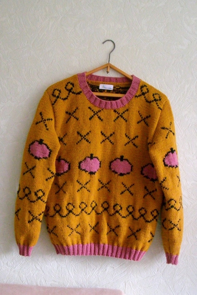 May include: A mustard yellow long-sleeve knit sweater featuring a repeating pattern of black cross stitches, black wavy lines, and pink pumpkin-shaped motifs. The garment has a pink ribbed crew neckline, cuffs, and hem, and a white brand label.